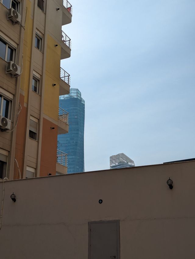 A view looking up at a beige apartment building on the left and a tall modern blue glass skyscraper in the distance against a blue sky.
