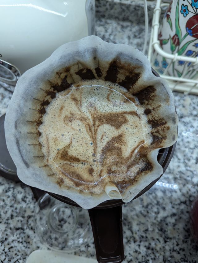 Top-down view of wet coffee grounds inside a white paper filter, showing a swirly pattern of foam and bubbles.